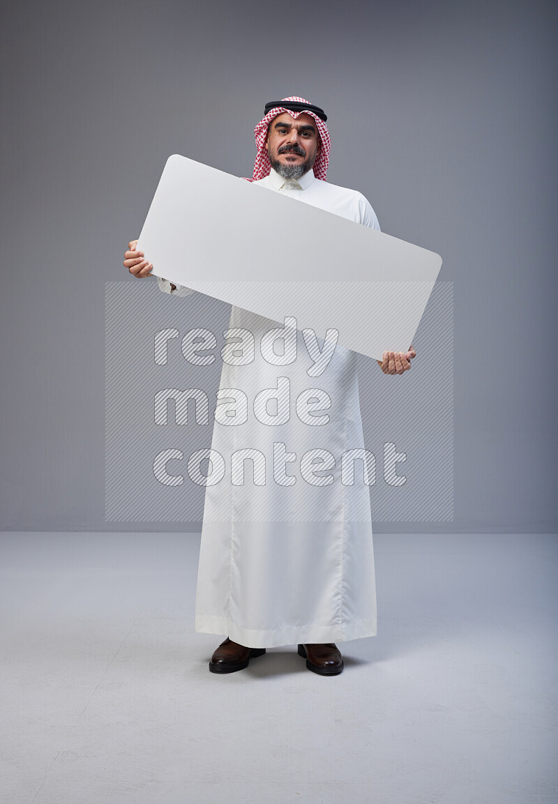 Saudi man Wearing Thob and red Shomag standing holding board on Gray background