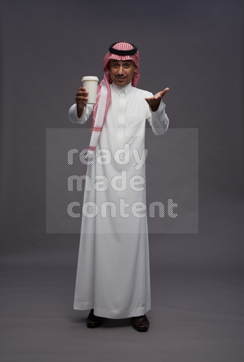 Saudi man wearing thob and shomag standing holding paper cup on gray background