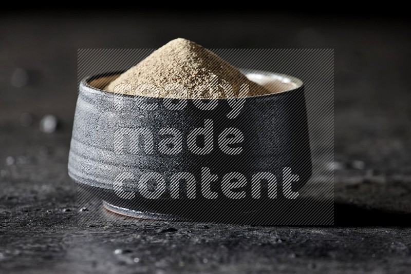 Black pottery bowl full of white pepper powder on textured black flooring