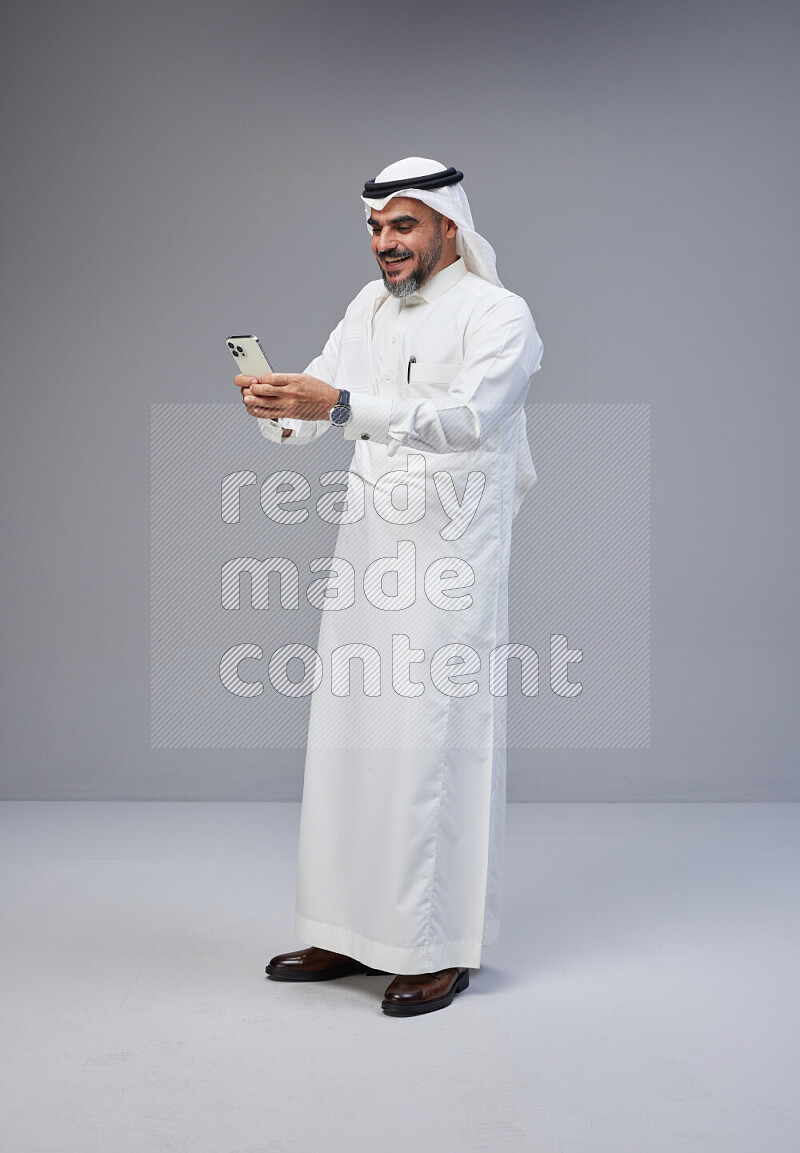 Saudi man Wearing Thob and white Shomag standing texting on phone on Gray background