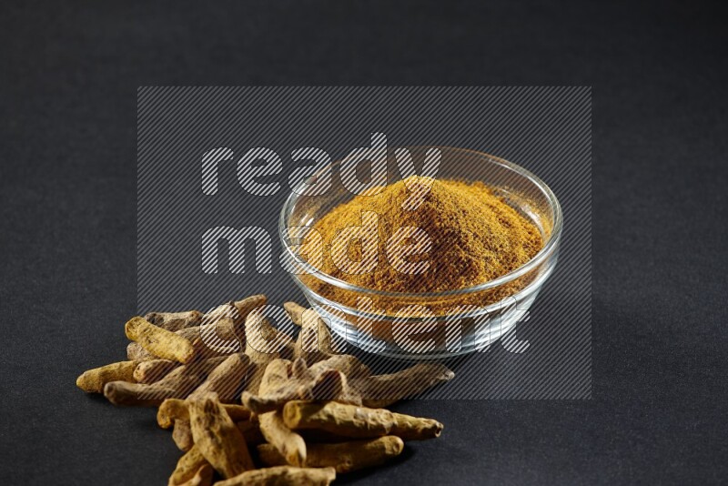 A glass bowl full of turmeric powder and dried whole fingers beside it on black flooring