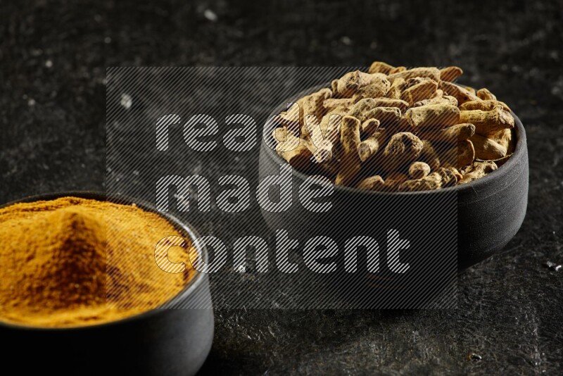2 pottery black bowls, one full of turmeric powder and the other full of dried turmeric whole fingers on a textured black flooring