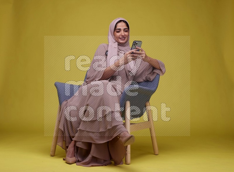 A Female Texting Sitting  on a Yellow Background wearing Brown Abaya with Hijab