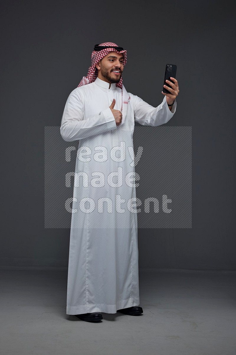 A Saudi man wearing Thobe and Shamgh taking selfie with his phone eye level on a gray background