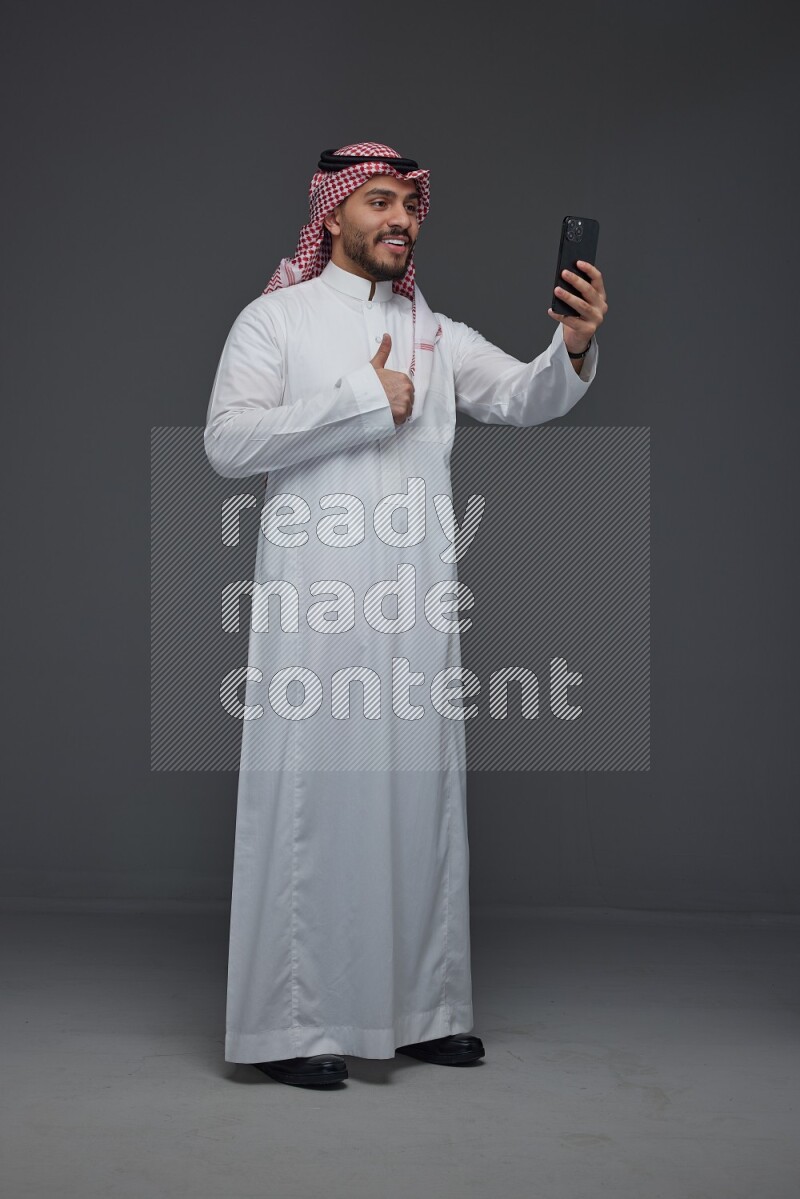 A Saudi man wearing Thobe and Shamgh taking selfie with his phone eye level on a gray background