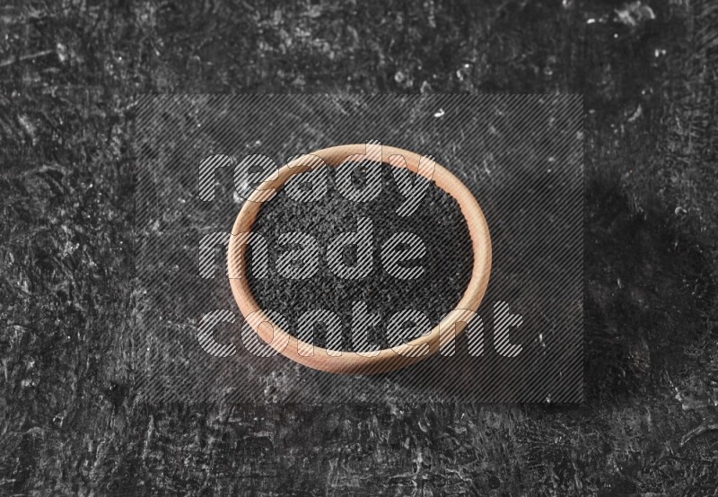 A wooden bowl full of black seeds on a textured black flooring