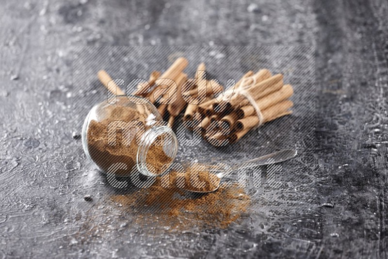 Herbal glass jar full cinnamon powder flipped and a metal spoon full of powder, cinnamon sticks stacked and bounded in the back on textured black background in different angles
