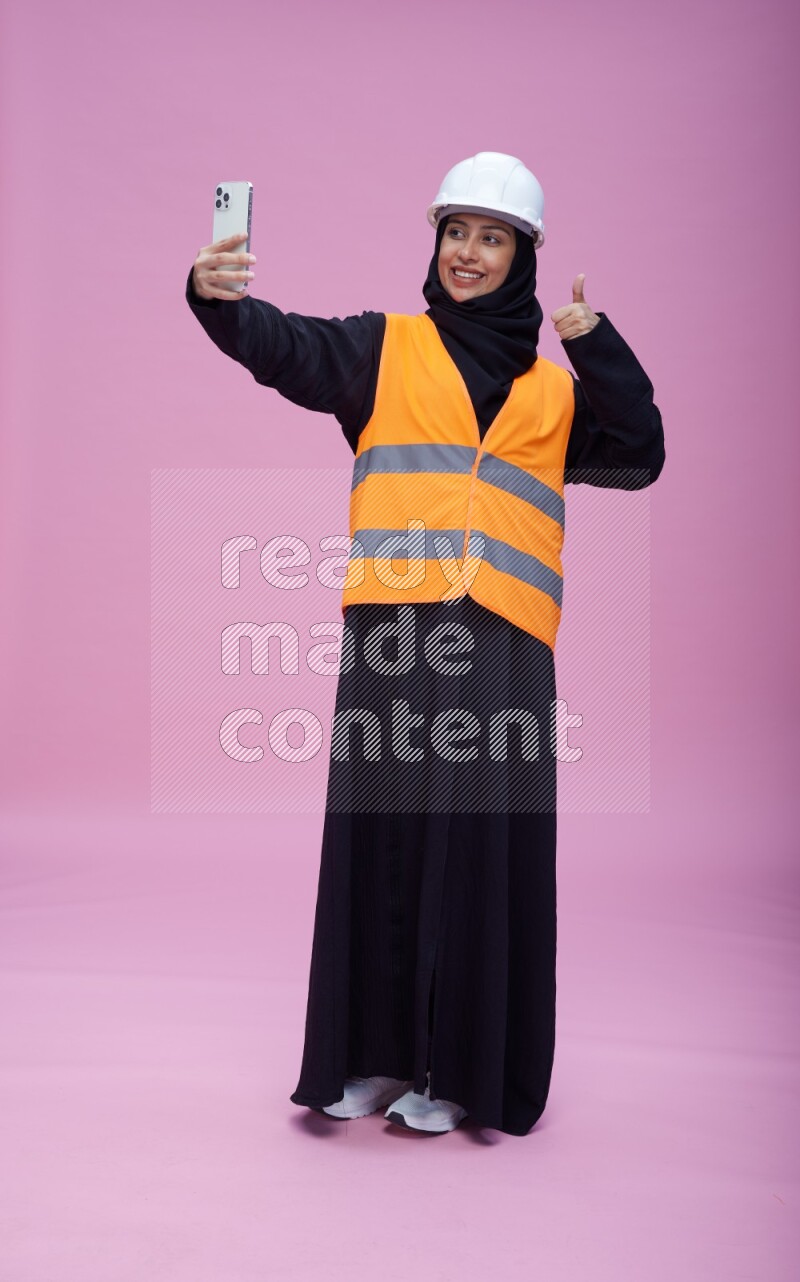 Saudi woman wearing Abaya with engineer vest and helmet standing taking selfie on pink background