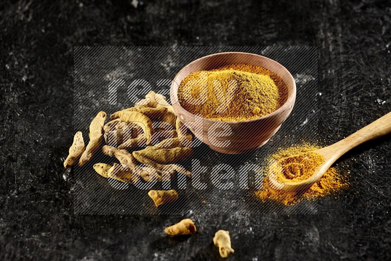 A wooden bowl and a wooden spoon full of turmeric powder with dried turmeric fingers on textured black flooring