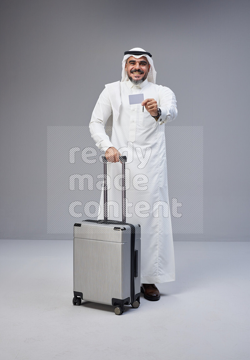 Saudi man wearing Thob and white Shomag standing holding Travel bag and ATM card on Gray background