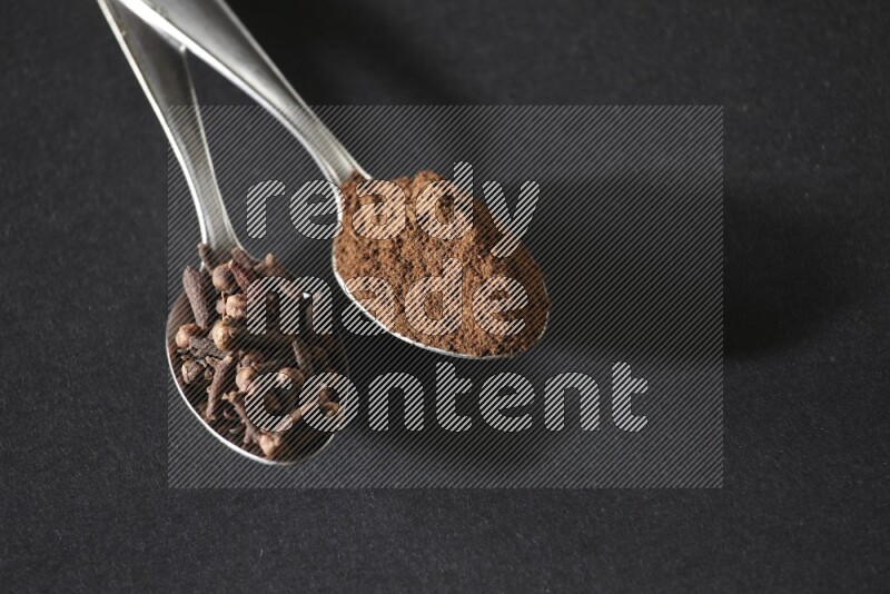 2 metal spoons, one full of cloves grains and the other full of cloves powder on a black flooring
