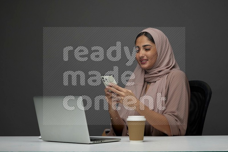 A Saudi woman Sitting on her desk Texting on a Gray Background wearing Brown Abaya with Hijab