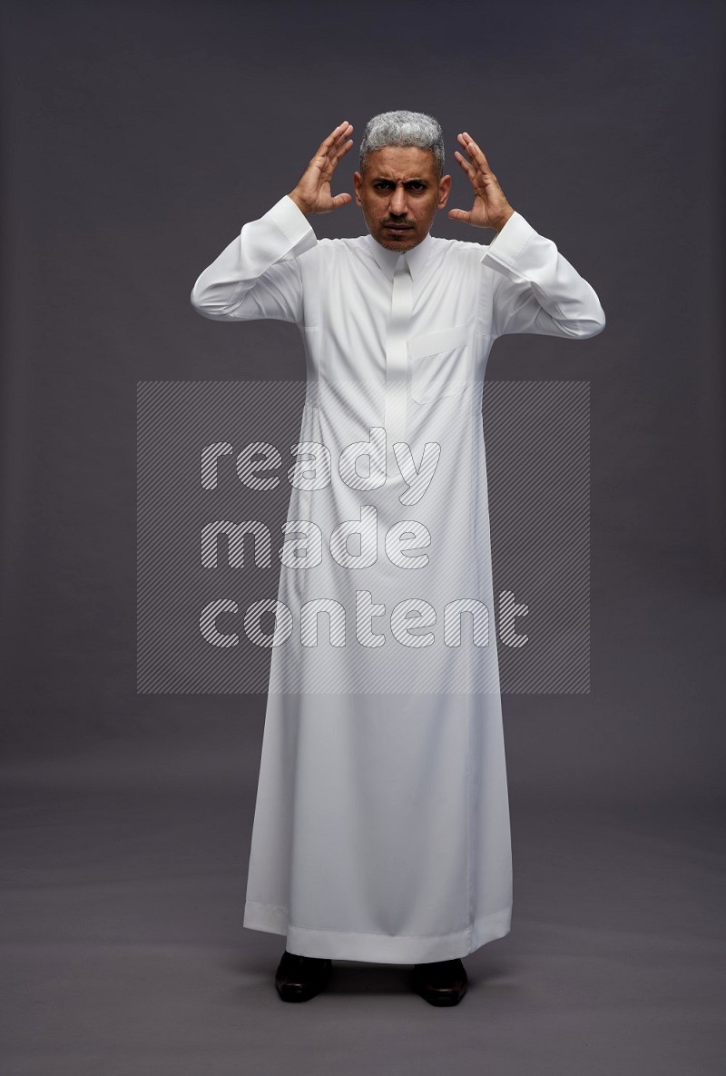 Saudi man wearing thob standing hands behind head on gray background