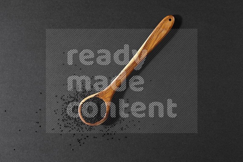 A wooden ladle full of black seeds and the seeds spread beside it on a black flooring