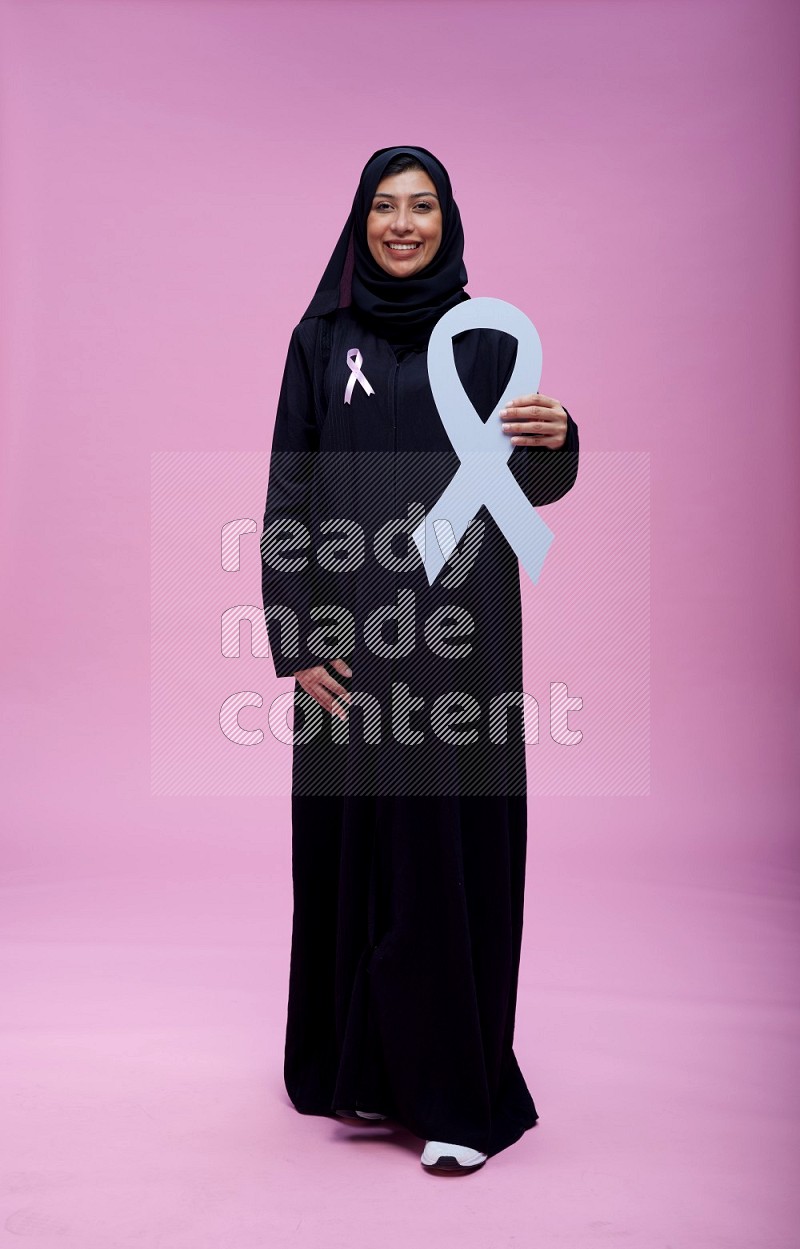 Saudi woman wearing pink ribbon on abaya standing holding awareness ribbon on plain pink background