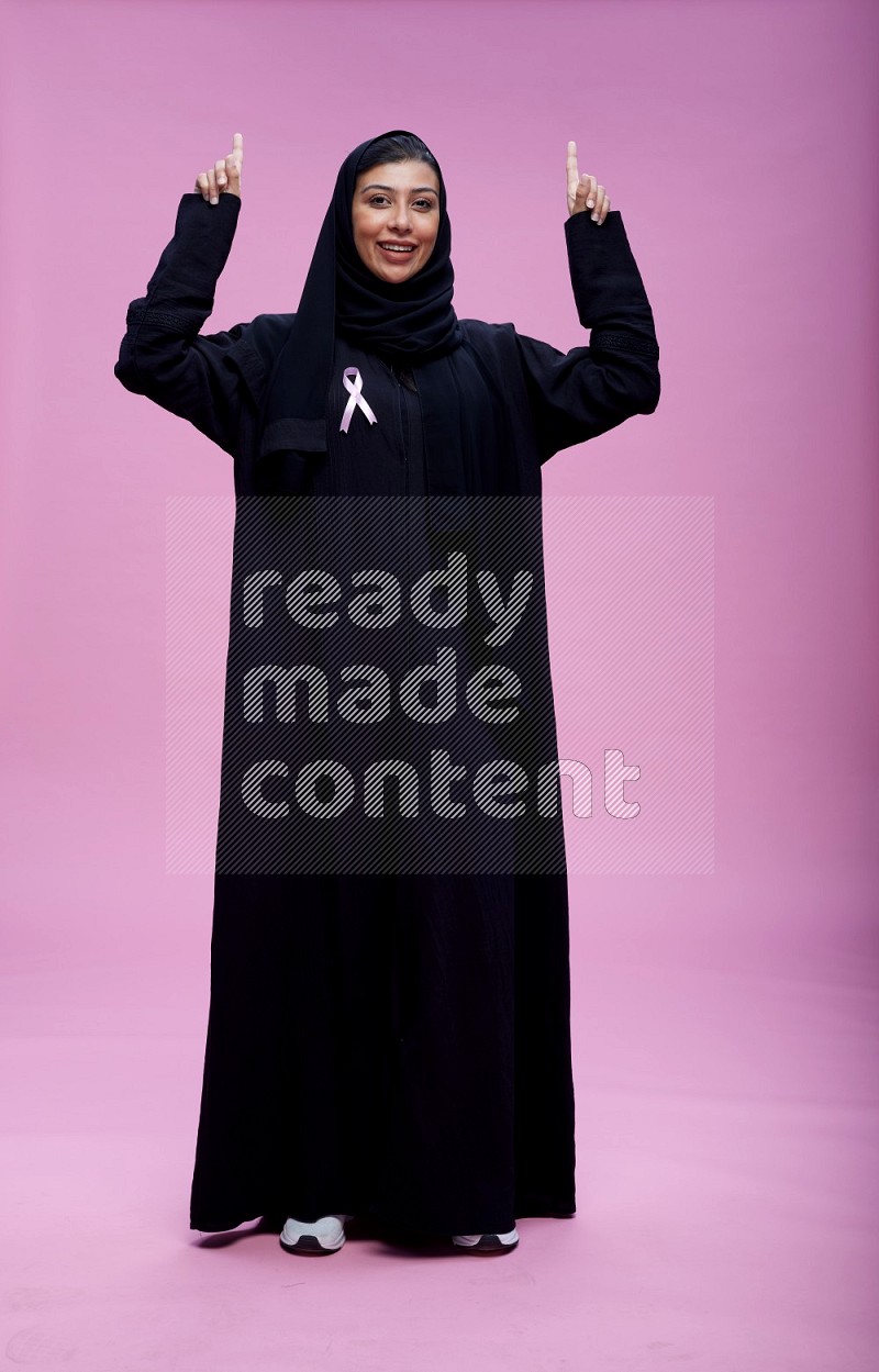 Saudi woman wearing pink ribbon on Abaya standing interacting with the camera on pink background