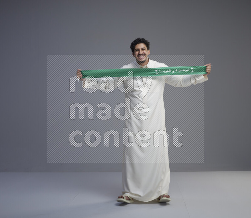 A Saudi man standing wearing thob holding Saudi flag scarf on gray background