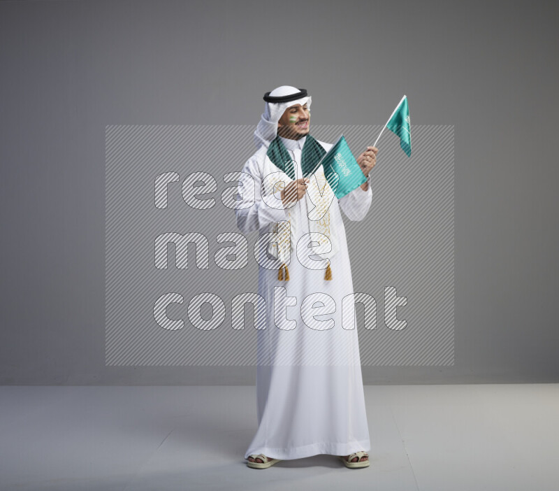 A saudi man standing wearing thob and white shomag with face painting and saudi flag scarf and holding small saudi flag on gray background