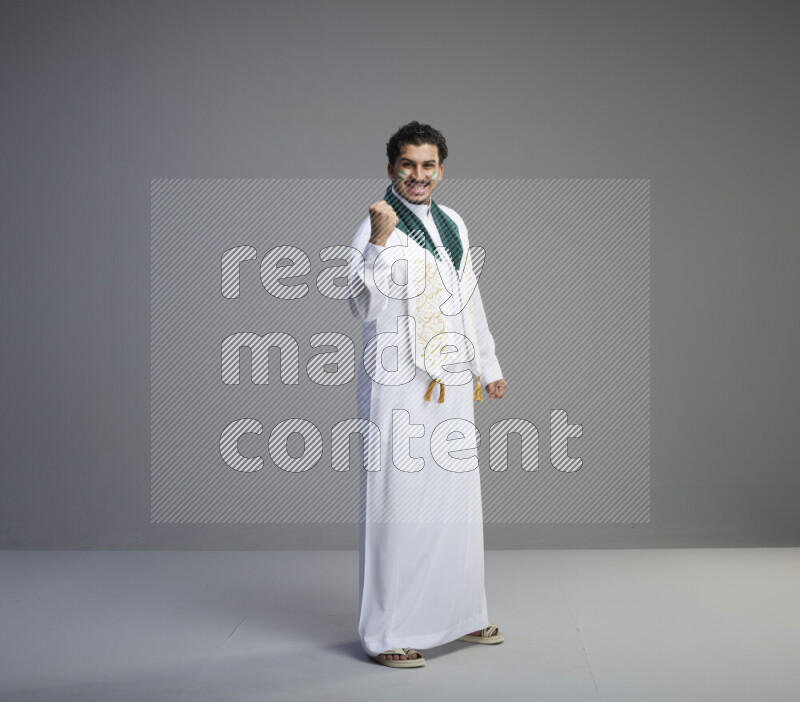 A Saudi man standing wearing thob and Saudi flag scarf with face painting on gray background