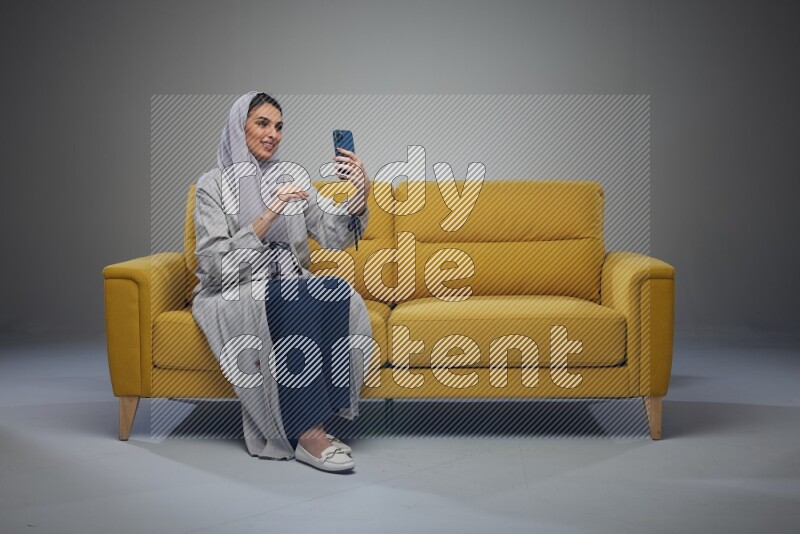 A Saudi woman wearing a light gray Abaya and head scarf sitting on a yellow sofa and making a video call eye level on a grey background