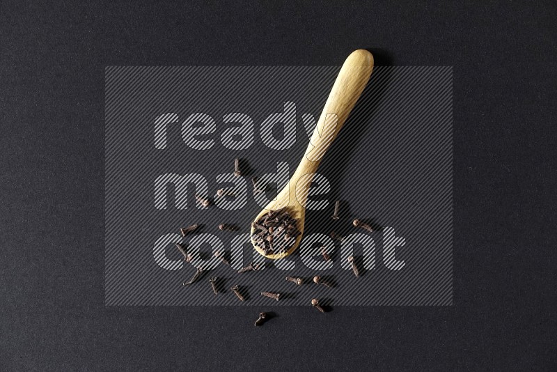 A wooden spoon full of cloves and some spreaded out of it on a black flooring