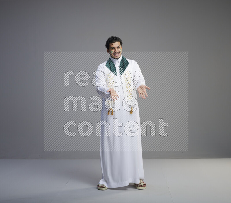 A Saudi man standing wearing thob and Saudi flag scarf with face painting on gray background