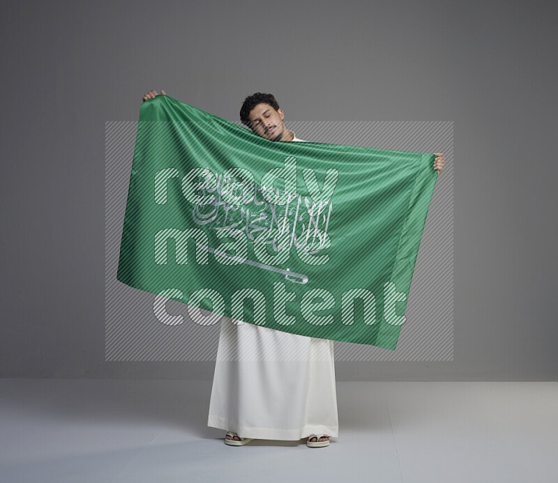 A saudi man standing wearing thob holding big saudi flag on gray background