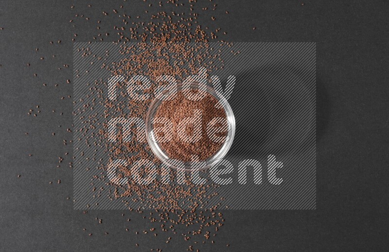 A glass bowl full of garden cress seeds surrounded by seeds on a black flooring