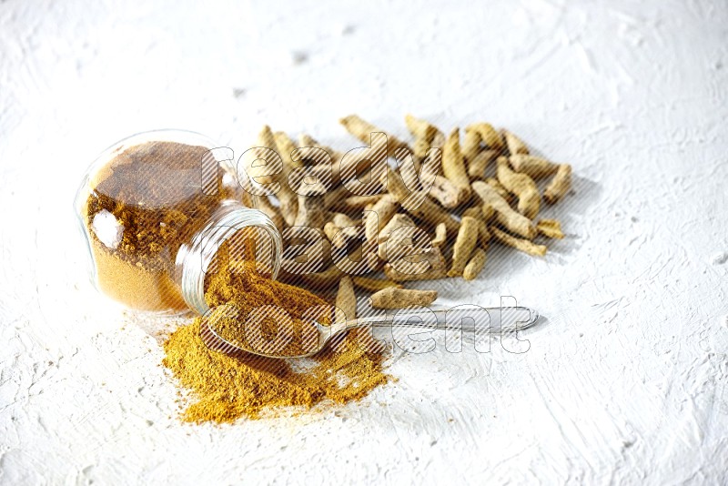 A flipped glass spice jar and metal spoon full of turmeric powder and powder spilled out of it with dried whole fingers on textured white flooring