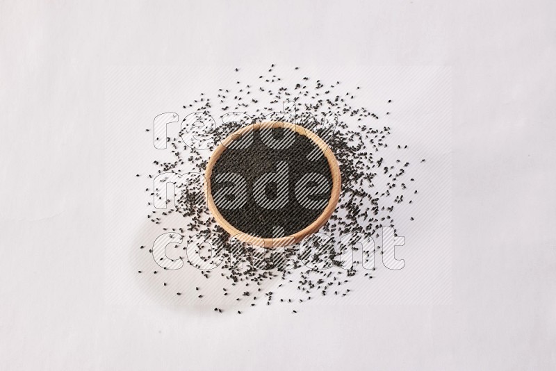A wooden bowl full of black seeds and more seeds spread on a white flooring