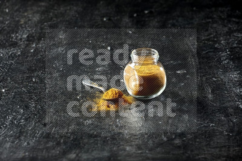 A glass spice jar and metal spoon full of turmeric powder on textured black flooring