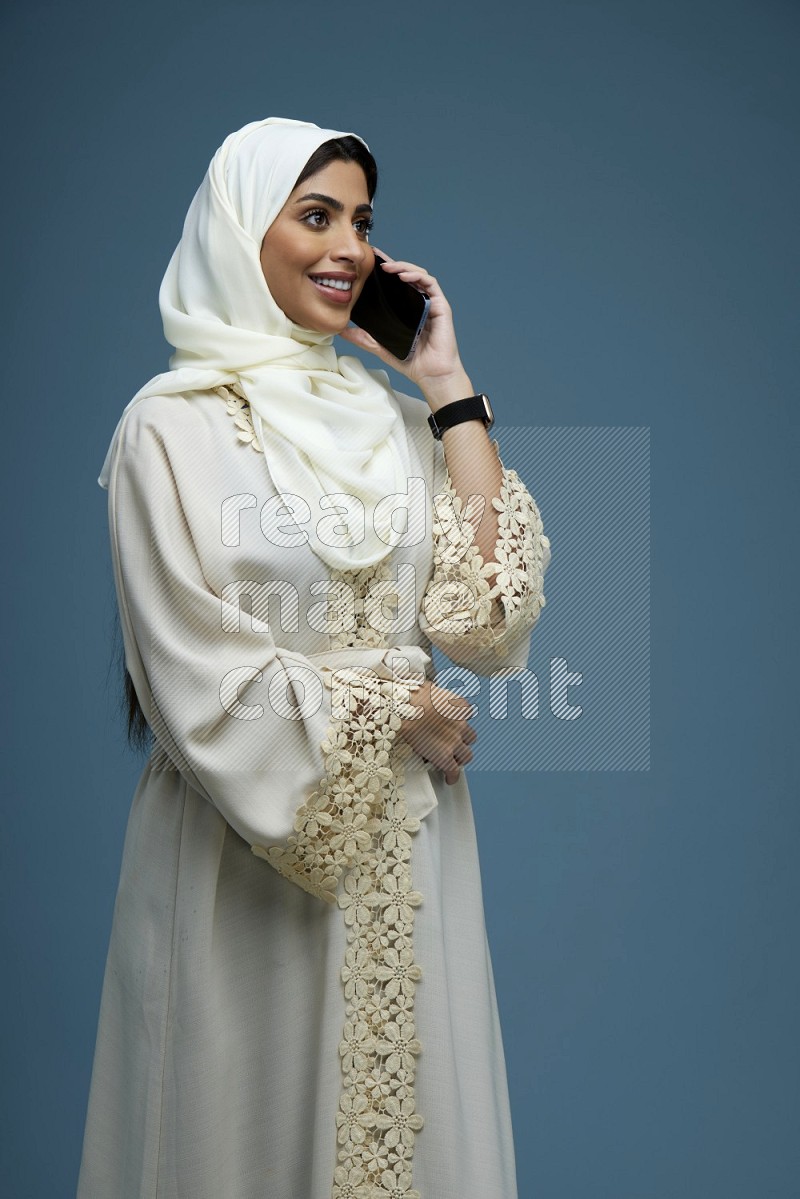 A Saudi woman having a call in a blue background wearing an off-white Abaya Hijab