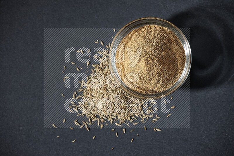 وعاء زجاجي ممتلئ ببودرة الكمون مع حبوب الكمون بجانبه على خلفية سوداء