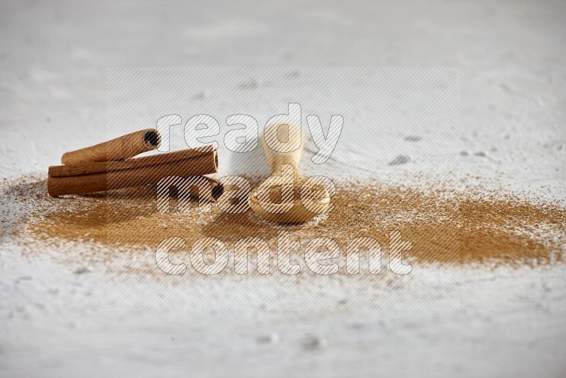 بودرة القرفة في ملعقة خشبية مع أعواد القرفة وبودرة متناثرة على الأرضية على خلفية بيضاء