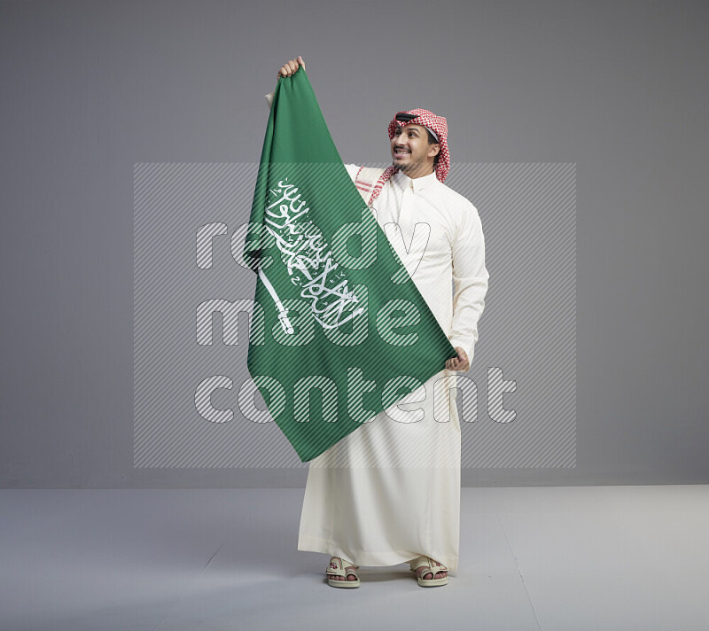 A saudi man standing wearing thob and red shomag holding big saudi flag on gray background