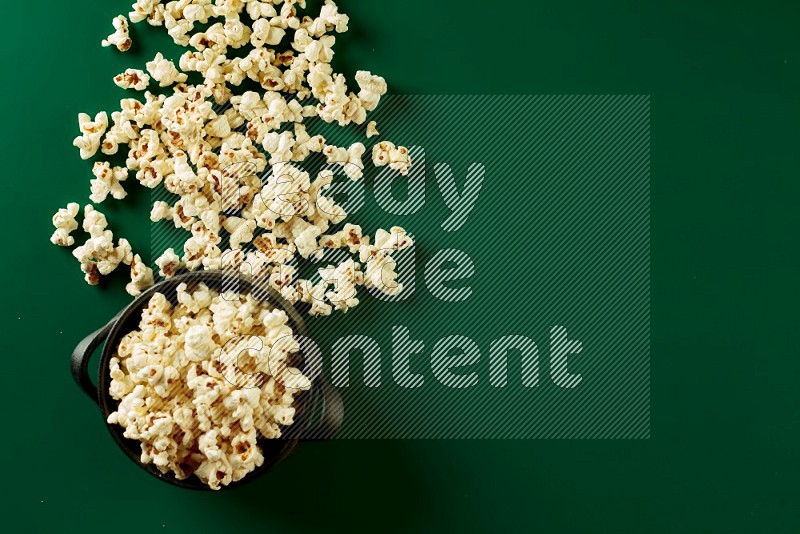 A black ceramic bowl full of popcorn with popcorn beside it on a green background in a top view shot