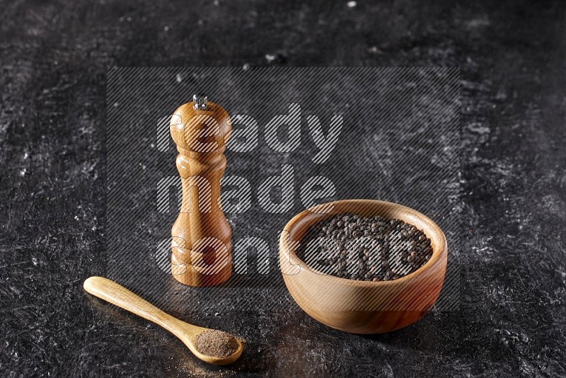 A wooden bowl full of black pepper and a wooden spoon full of black pepper powder and a wooden grinder on a textured black flooring