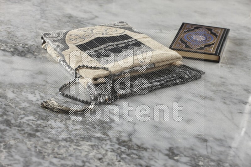 A folded prayer mat with different elements such as quran and prayer beads on grey marble background