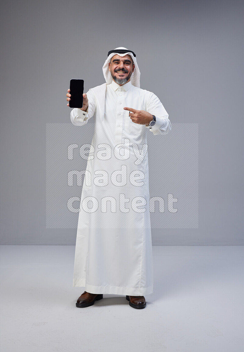 Saudi man Wearing Thob and white Shomag standing showing phone to camera on Gray background