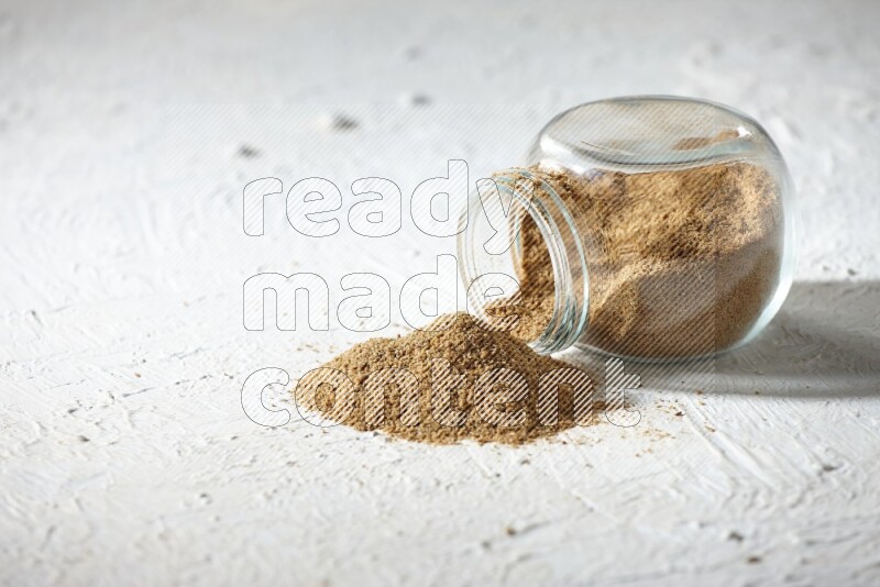 A flipped glass spice jar full of cumin powder and powder spilled out on textured white flooring