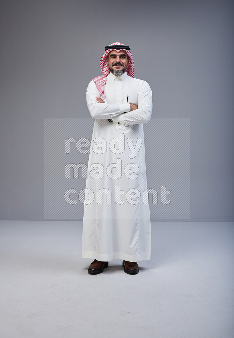 Saudi man Wearing Thob and red Shomag standing with crossed arms on Gray background
