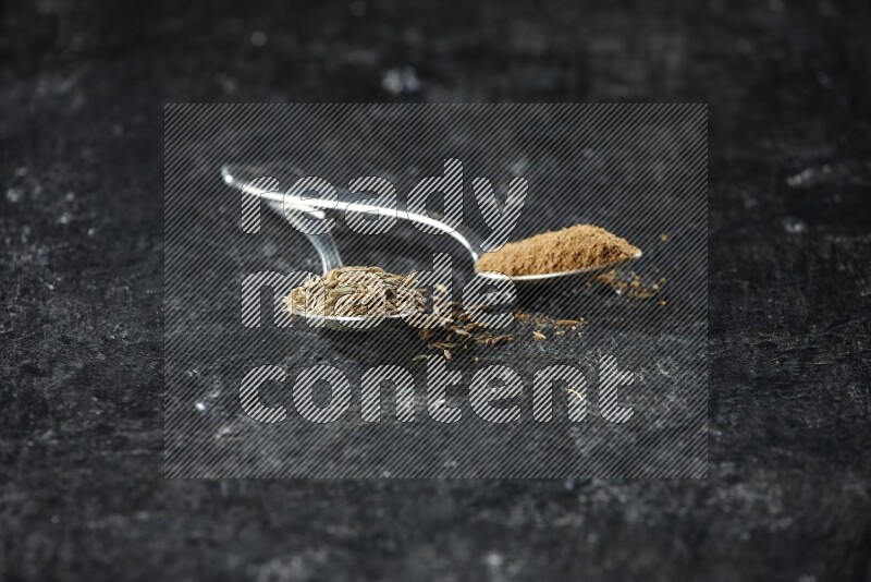 ملعقتان معدنيتان مملوءتان بحبوب الكمون وبودرة الكمون على أرضية سوداء