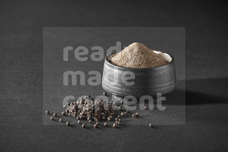 A black pottery bowl full of black pepper powder and black pepper beads on the floor on black flooring