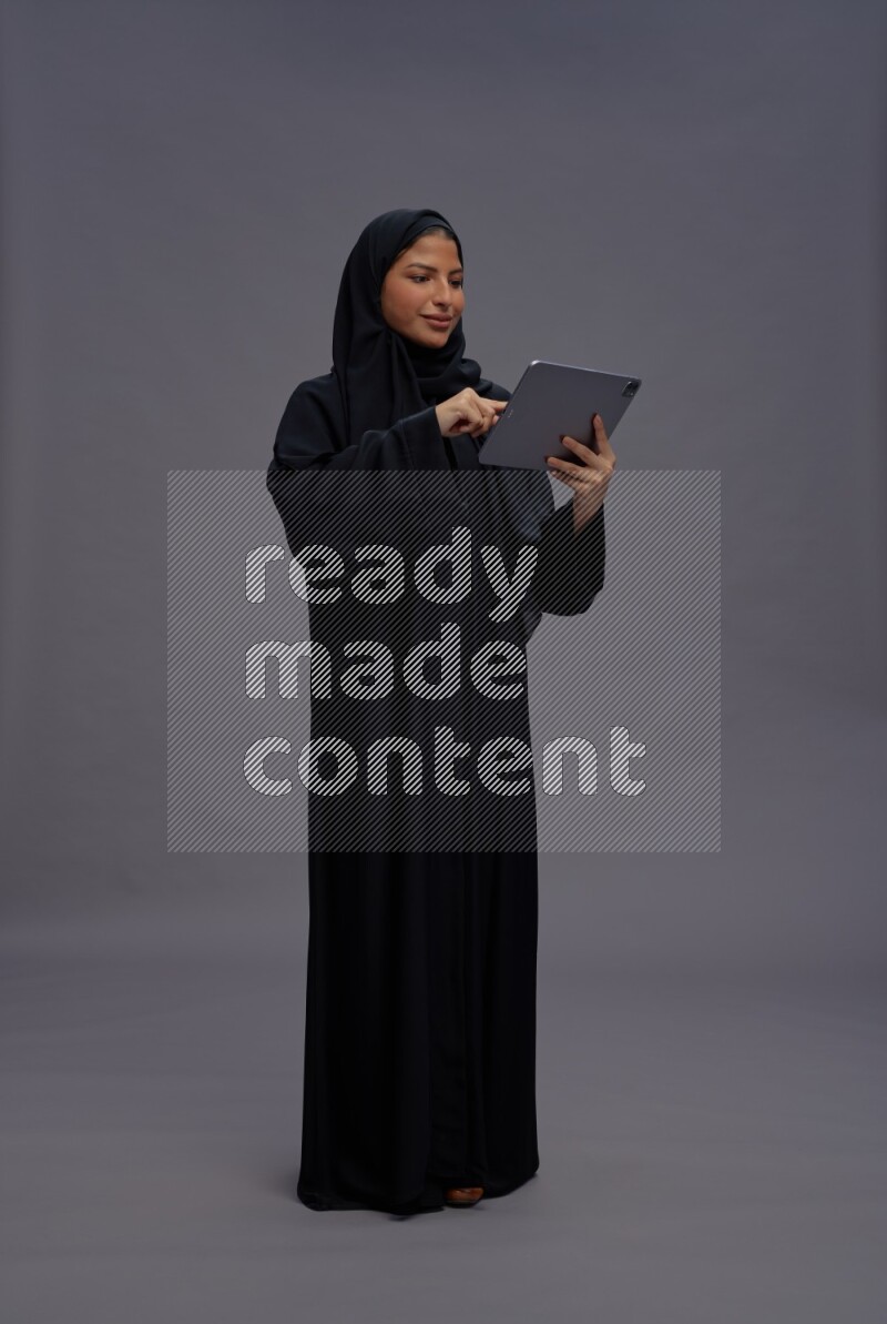 Saudi woman wearing Abaya standing working on tablet on gray background