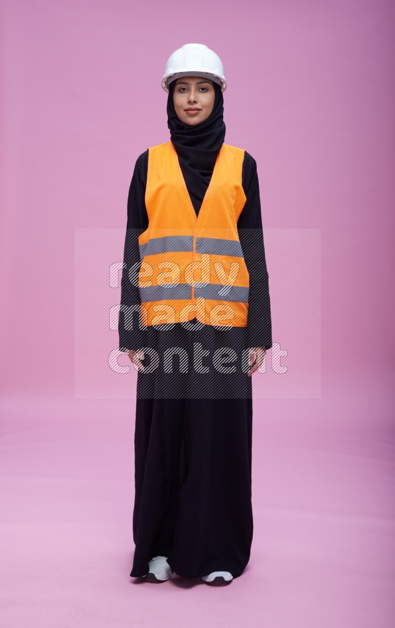 Saudi woman wearing Abaya with engineer vest and helmet standing interacting with the camera on pink background