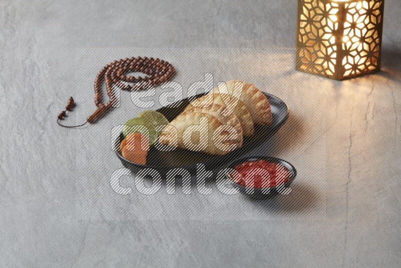 Four fried sambosas in an oval shaped black plate, beside a cut cheese sambosa, a brown misbaha and a golden lantern on a gray background