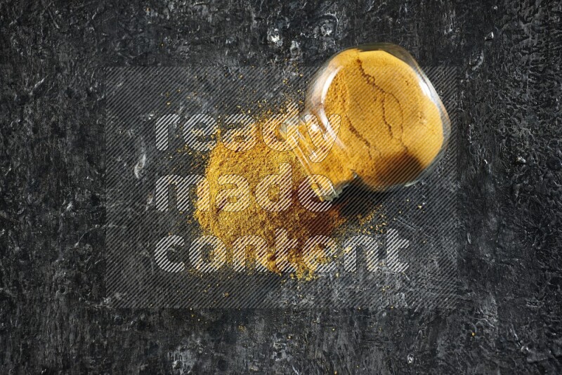 وعاء زجاجي للتوابل مقلوب وممتلئ ببودرة الكركم والبودرة تناثرت منه على خلفية سوداء