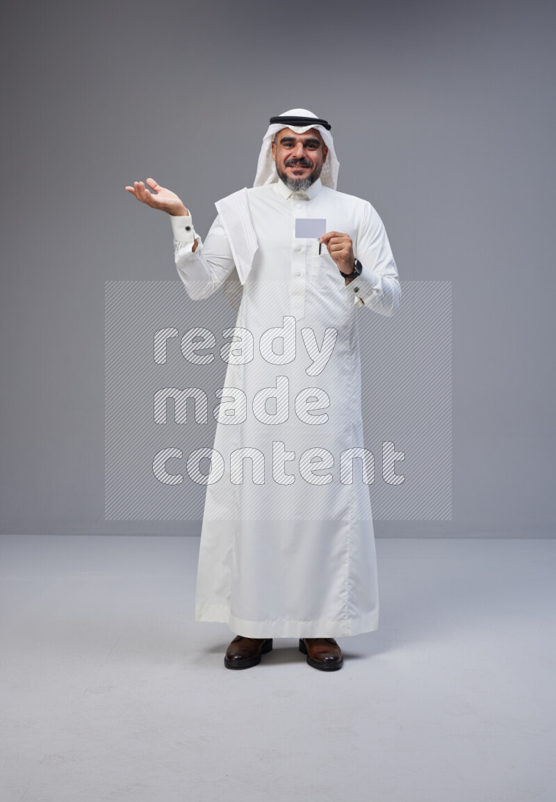 Saudi man Wearing Thob and white Shomag standing holding ATM card on Gray background