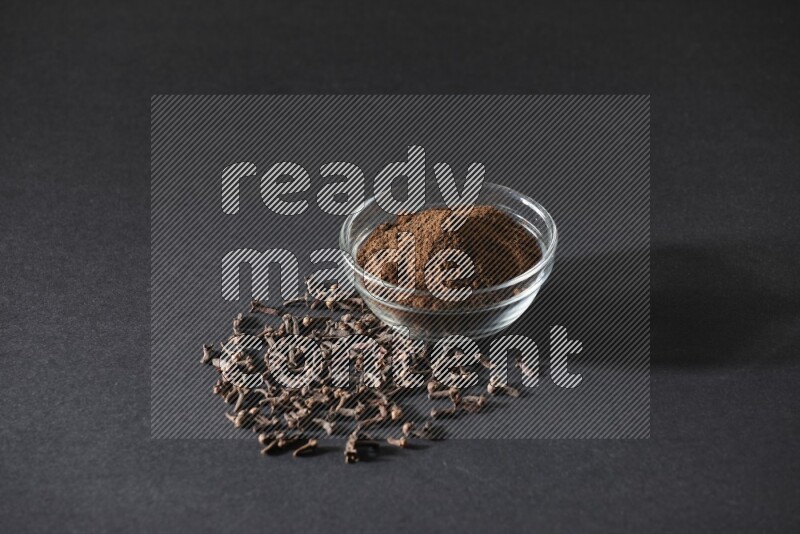 A glass bowl full of cloves powder with cloves grains spread on black flooring