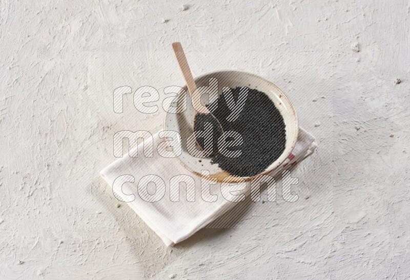 A multicolored pottery plate full of black seeds with a wooden spoon full of the seeds on a napkin on a textured white flooring